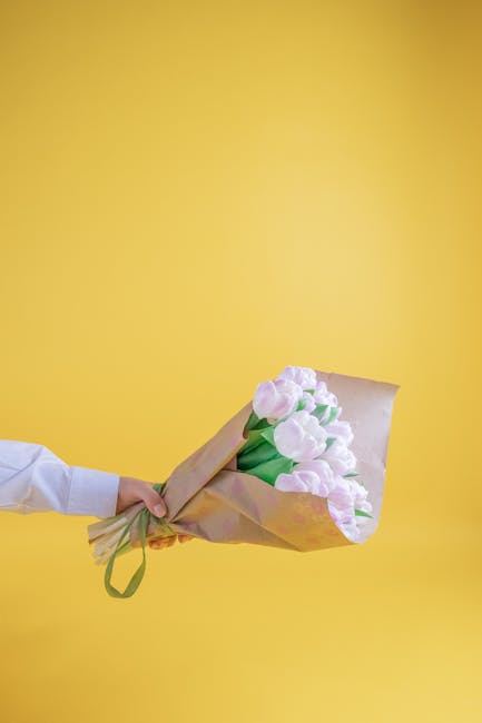 A person wearing a white shirt holds a bouquet of fresh pink and white tulips wrapped in light brown paper with a green ribbon, extending the floral arrangement toward the camera against a solid yello