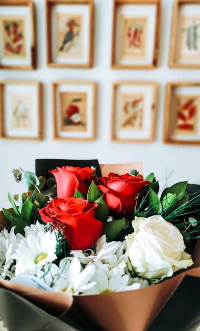 A close-up view of a floral arrangement featuring vibrant red roses and white daisies, expertly wrapped in soft pink tissue paper with green foliage accents. The bouquet is positioned in the foregroun