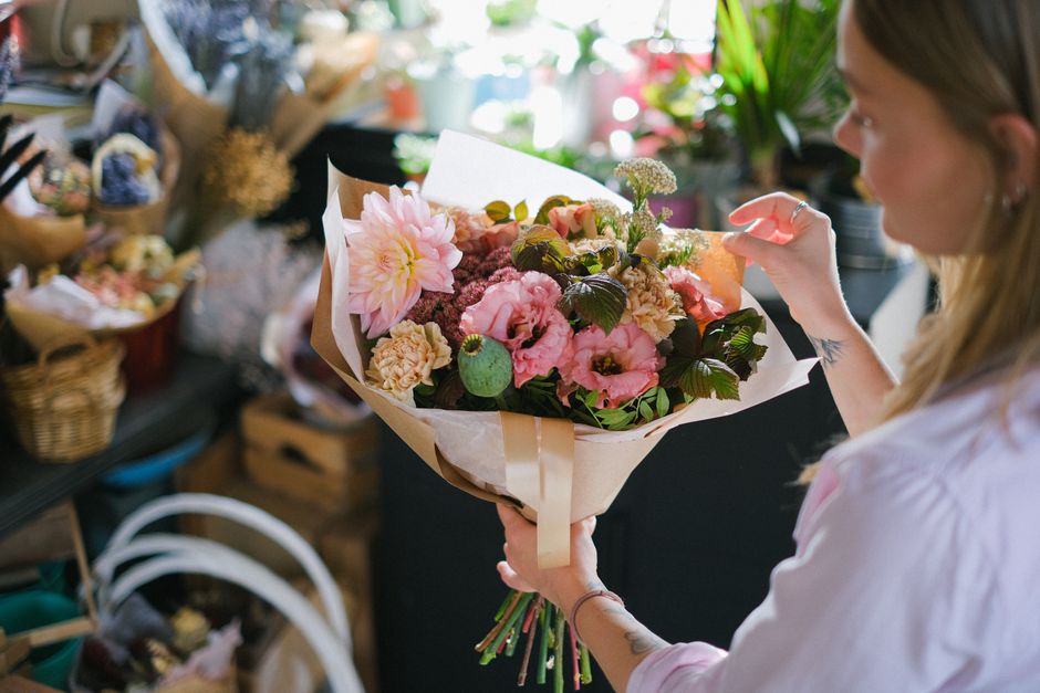 A woman holding a bouquet of fresh flowers featuring pink dahlias, lisianthus, and carnations wrapped in light brown and white paper with a satin ribbon, standing in a flower shop with various floral 