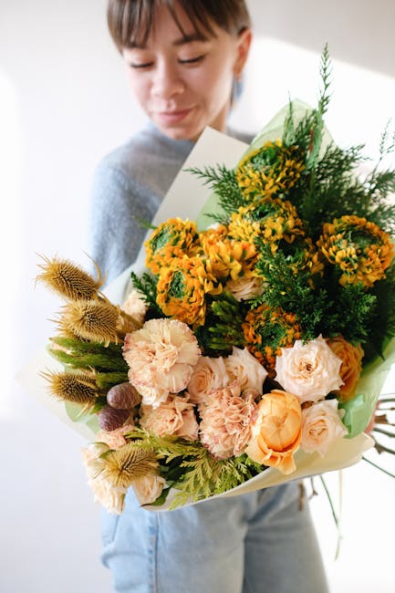 A young person holding a large floral gift bouquet featuring a mix of fresh, vibrant flowers. The arrangement includes peachy roses, cheerful yellow mimosa-like blossoms, and soft pink carnations, com